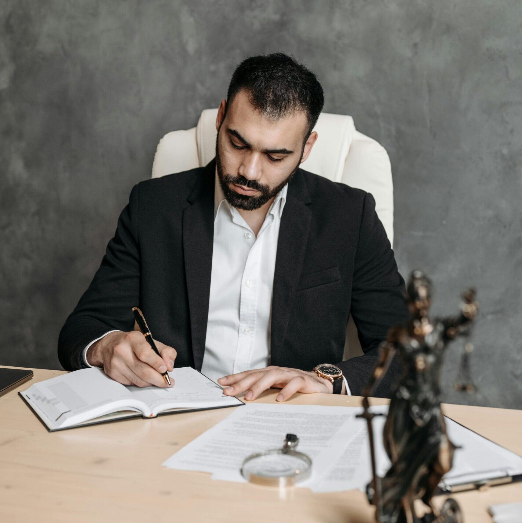 Professional lawyer in a suit taking notes at a desk with legal documents and statue.